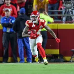 Skyy Moore, Wide Receiver der Kansas City Chiefs, in roter Uniform und weißem Helm, läuft während eines Spiels mit dem Football auf einem grünen Feld. Fans und Mitarbeiter, einige in Gelb und Blau, beobachten von der Tribüne aus hinter einer roten Absperrung, wie er nach rechts läuft. Diese Beschreibung wurde mit der FootballR KI automatisch generiert.