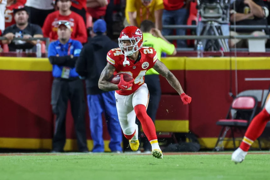 Skyy Moore, Wide Receiver der Kansas City Chiefs, in roter Uniform und weißem Helm, läuft während eines Spiels mit dem Football auf einem grünen Feld. Fans und Mitarbeiter, einige in Gelb und Blau, beobachten von der Tribüne aus hinter einer roten Absperrung, wie er nach rechts läuft. Diese Beschreibung wurde mit der FootballR KI automatisch generiert.