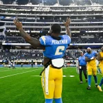 Ein Football-Spieler, Denzel Perryman, trägt ein blau-gelbes Trikot mit der Aufschrift "Perryman 6" auf dem Rücken und hebt beide Arme in Richtung der Zuschauer im Stadion. Andere Mannschaftskameraden und Mitarbeiter laufen hinter ihm auf dem grünen Feld, die Sitze sind im Hintergrund zu sehen. Diese Beschreibung wurde mit der FootballR KI automatisch generiert.