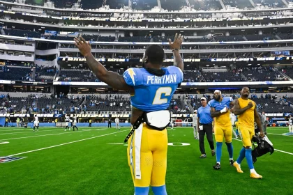 Ein Football-Spieler, Denzel Perryman, trägt ein blau-gelbes Trikot mit der Aufschrift "Perryman 6" auf dem Rücken und hebt beide Arme in Richtung der Zuschauer im Stadion. Andere Mannschaftskameraden und Mitarbeiter laufen hinter ihm auf dem grünen Feld, die Sitze sind im Hintergrund zu sehen. Diese Beschreibung wurde mit der FootballR KI automatisch generiert.