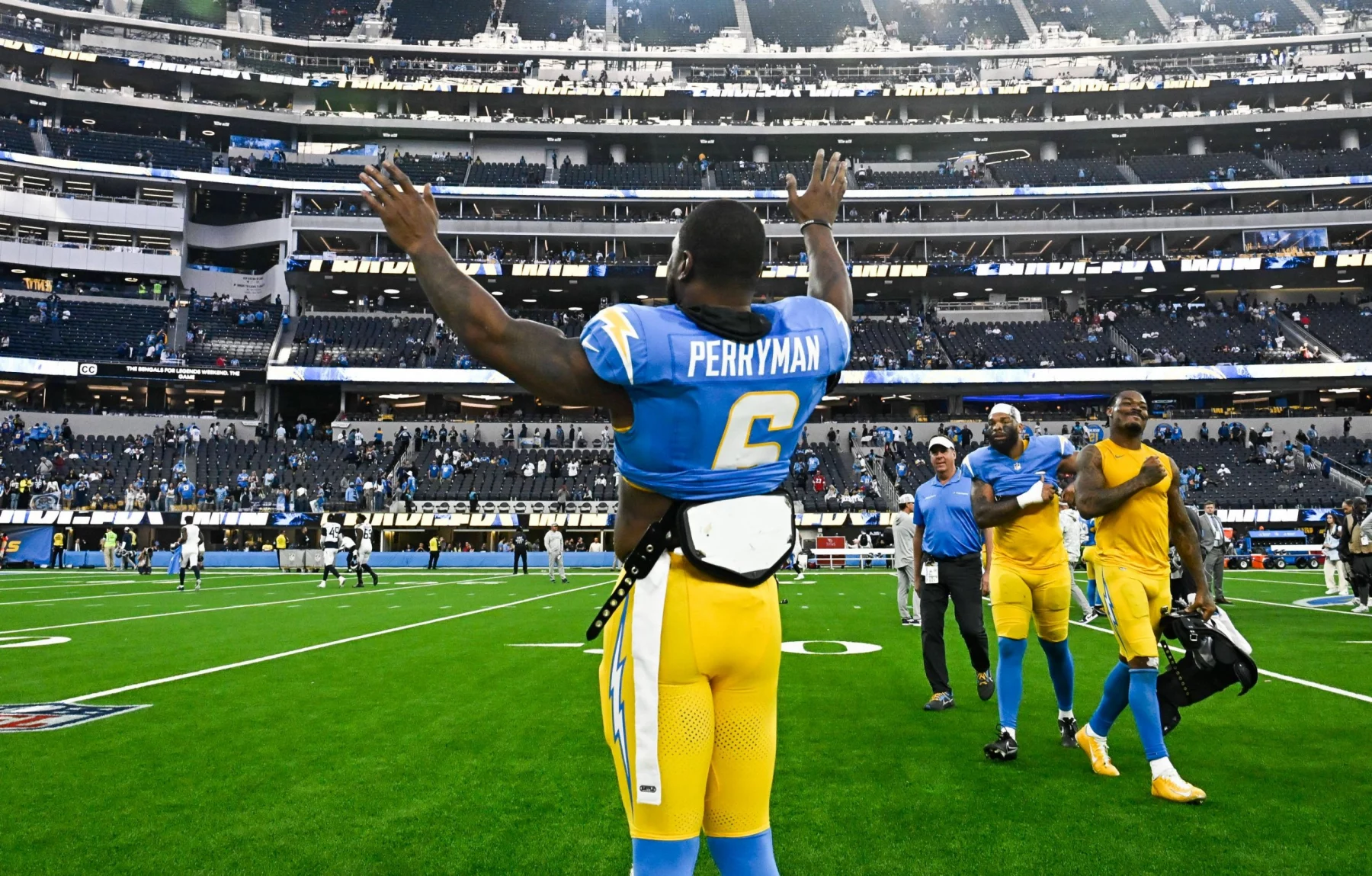 Ein Football-Spieler, Denzel Perryman, trägt ein blau-gelbes Trikot mit der Aufschrift "Perryman 6" auf dem Rücken und hebt beide Arme in Richtung der Zuschauer im Stadion. Andere Mannschaftskameraden und Mitarbeiter laufen hinter ihm auf dem grünen Feld, die Sitze sind im Hintergrund zu sehen. Diese Beschreibung wurde mit der FootballR KI automatisch generiert.