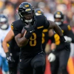 Brian Robinson Jr., der eine schwarze Uniform mit der Nummer 8 und einen schwarzen Helm trägt, läuft während eines Spiels mit dem Ball. Im Hintergrund sind andere Spieler und unscharfe Zuschauer zu sehen, die das schnelle Geschehen auf dem Spielfeld festhalten. Diese Beschreibung wurde mit der FootballR KI automatisch generiert.