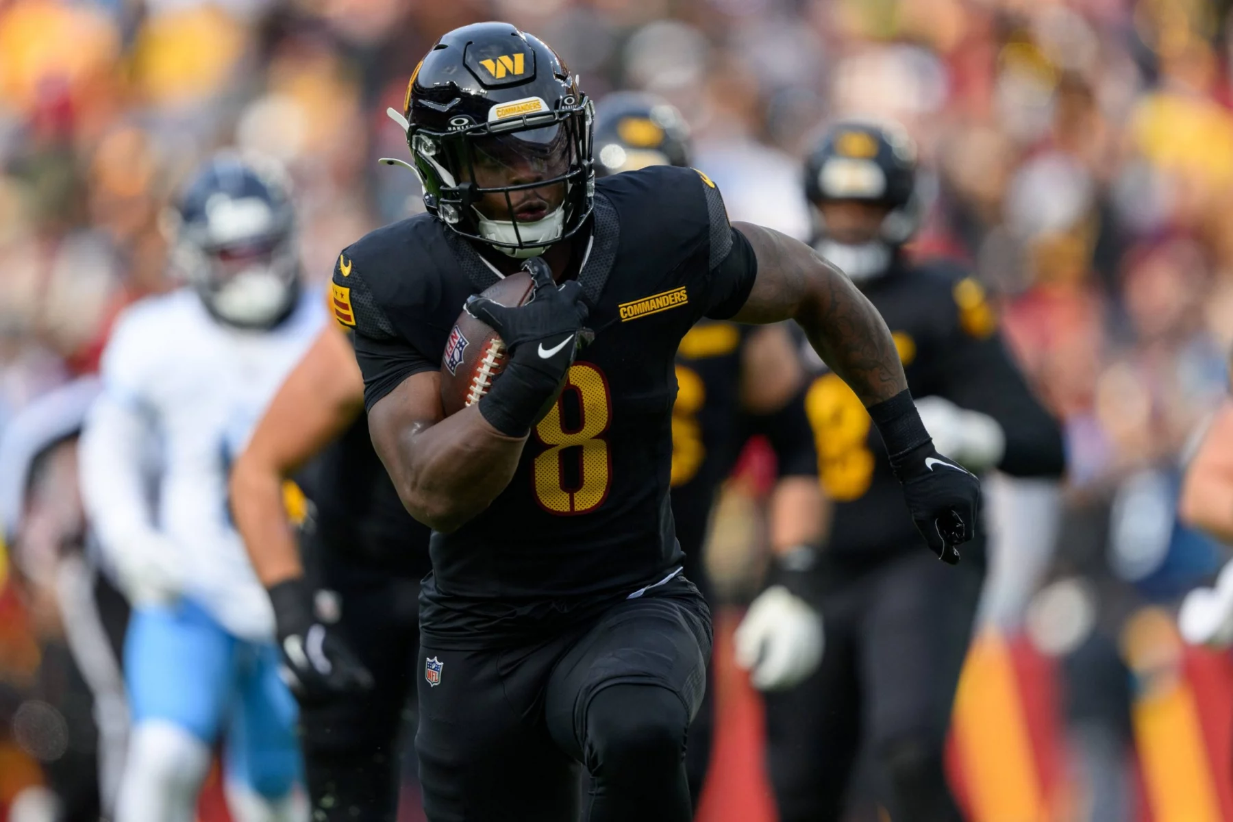 Brian Robinson Jr., der eine schwarze Uniform mit der Nummer 8 und einen schwarzen Helm trägt, läuft während eines Spiels mit dem Ball. Im Hintergrund sind andere Spieler und unscharfe Zuschauer zu sehen, die das schnelle Geschehen auf dem Spielfeld festhalten. Diese Beschreibung wurde mit der FootballR KI automatisch generiert.