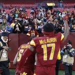 Ein Footballspieler in einem burgunderroten und gelben Trikot mit der Aufschrift "McLaurin 17" auf dem Rücken - Terry McLaurin - hebt seinen Arm in Richtung der jubelnden Fans auf den Tribünen. Fotografen halten den Moment fest, während die in Winterkleidung gekleideten Fans hinter einer Absperrung feiern. Diese Beschreibung wurde mit der FootballR KI automatisch generiert.