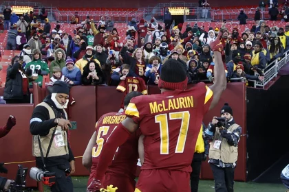 Ein Footballspieler in einem burgunderroten und gelben Trikot mit der Aufschrift "McLaurin 17" auf dem Rücken - Terry McLaurin - hebt seinen Arm in Richtung der jubelnden Fans auf den Tribünen. Fotografen halten den Moment fest, während die in Winterkleidung gekleideten Fans hinter einer Absperrung feiern. Diese Beschreibung wurde mit der FootballR KI automatisch generiert.