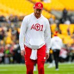Carson Wentz, ein Footballspieler mit roter Mütze, weißem "AO1"-Löwen-Kapuzenpulli und roten Shorts, steht auf einem Feld mit gelben Stadionsitzen und unscharfen Menschen im Hintergrund. Das Wetter ist klar und die Stimmung ist locker. Diese Beschreibung wurde mit der FootballR KI automatisch generiert.