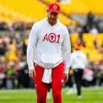 Carson Wentz, ein Footballspieler mit roter Mütze, weißem "AO1"-Löwen-Kapuzenpulli und roten Shorts, steht auf einem Feld mit gelben Stadionsitzen und unscharfen Menschen im Hintergrund. Das Wetter ist klar und die Stimmung ist locker. Diese Beschreibung wurde mit der FootballR KI automatisch generiert.