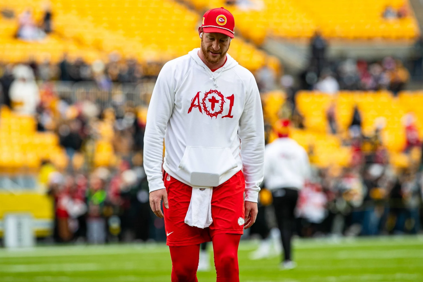 Carson Wentz, ein Footballspieler mit roter Mütze, weißem "AO1"-Löwen-Kapuzenpulli und roten Shorts, steht auf einem Feld mit gelben Stadionsitzen und unscharfen Menschen im Hintergrund. Das Wetter ist klar und die Stimmung ist locker. Diese Beschreibung wurde mit der FootballR KI automatisch generiert.