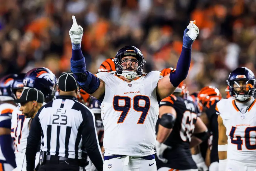 Zach Allen, Footballspieler der Denver Broncos (Nr. 99), jubelt mit erhobenen Armen und nach oben zeigenden Zeigefingern während eines Spiels, umgeben von Teamkollegen und Spielern der Cincinnati Bengals. Ein Schiedsrichter steht in der Nähe, während das Stadionpublikum im Hintergrund verschwommen ist. Diese Beschreibung wurde mit der FootballR KI automatisch generiert.