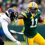 Woche 14 - Der Wide Receiver der Green Bay Packers, Jayden Reed, trägt den Football und hält einen Verteidiger der Chicago Bears während eines Spiels fest im Griff. Reed trägt ein grün-gelbes Trikot, während der Spieler der Bears ein weißes Trikot mit marineblauen und orangefarbenen Details trägt. Der Hintergrund ist unscharf. Diese Beschreibung wurde mit der FootballR KI automatisch generiert.