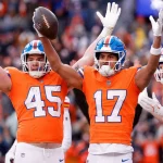 Drei Footballspieler in orangefarbenen Trikots feiern auf dem Spielfeld. Devaughn Vele, Nummer 17, reißt seine Arme zum Sieg hoch, während sein Teamkollege 45 einen Football hält. Der Hintergrund zeigt eine unscharfe Menge von Fans im Stadion. Diese Beschreibung wurde mit der FootballR KI automatisch generiert.