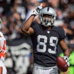 Ein Footballspieler, Amari Cooper, in einer schwarzen Raiders-Uniform mit der Nummer 89 hält einen Football in der einen Hand und zeigt mit der anderen nach oben. Er trägt einen silbernen Helm und ein Visier, während Spieler und Fans der Browns den Hintergrund füllen. Diese Beschreibung wurde mit der FootballR KI automatisch generiert.