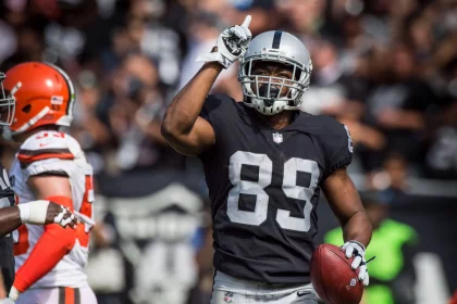 Ein Footballspieler, Amari Cooper, in einer schwarzen Raiders-Uniform mit der Nummer 89 hält einen Football in der einen Hand und zeigt mit der anderen nach oben. Er trägt einen silbernen Helm und ein Visier, während Spieler und Fans der Browns den Hintergrund füllen. Diese Beschreibung wurde mit der FootballR KI automatisch generiert.