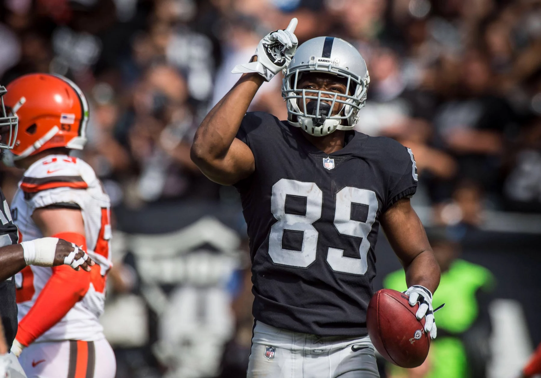 Ein Footballspieler, Amari Cooper, in einer schwarzen Raiders-Uniform mit der Nummer 89 hält einen Football in der einen Hand und zeigt mit der anderen nach oben. Er trägt einen silbernen Helm und ein Visier, während Spieler und Fans der Browns den Hintergrund füllen. Diese Beschreibung wurde mit der FootballR KI automatisch generiert.