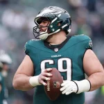 Ein Footballspieler in einem grünen Philadelphia Eagles Trikot und Helm, Nummer 69, hält einen Football in beiden Händen. Er trägt weiße Handschuhe und schaut zur Seite, als Landon Dickerson nach seiner Verletzung zurückkehrt. Das Stadion ist hinter ihm verschwommen. Diese Beschreibung wurde mit der FootballR KI automatisch generiert.