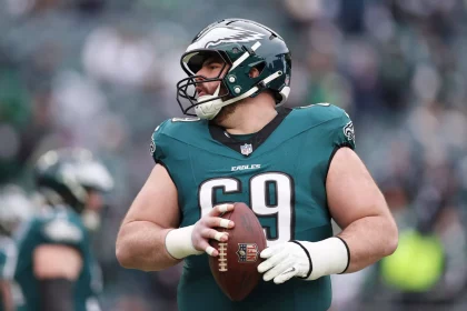 Ein Footballspieler in einem grünen Philadelphia Eagles Trikot und Helm, Nummer 69, hält einen Football in beiden Händen. Er trägt weiße Handschuhe und schaut zur Seite, als Landon Dickerson nach seiner Verletzung zurückkehrt. Das Stadion ist hinter ihm verschwommen. Diese Beschreibung wurde mit der FootballR KI automatisch generiert.