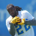 Ein Footballspieler mit Dreadlocks, der Najee Harris ähnelt, trägt ein weißes Trikot mit der Nummer 22 und gelbe Handschuhe und bereitet sich darauf vor, einen Football beim Training zu fangen. Der blaue, leicht verschwommene Hintergrund hebt die Aktion des Sportlers hervor. Diese Beschreibung wurde mit der FootballR KI automatisch generiert.