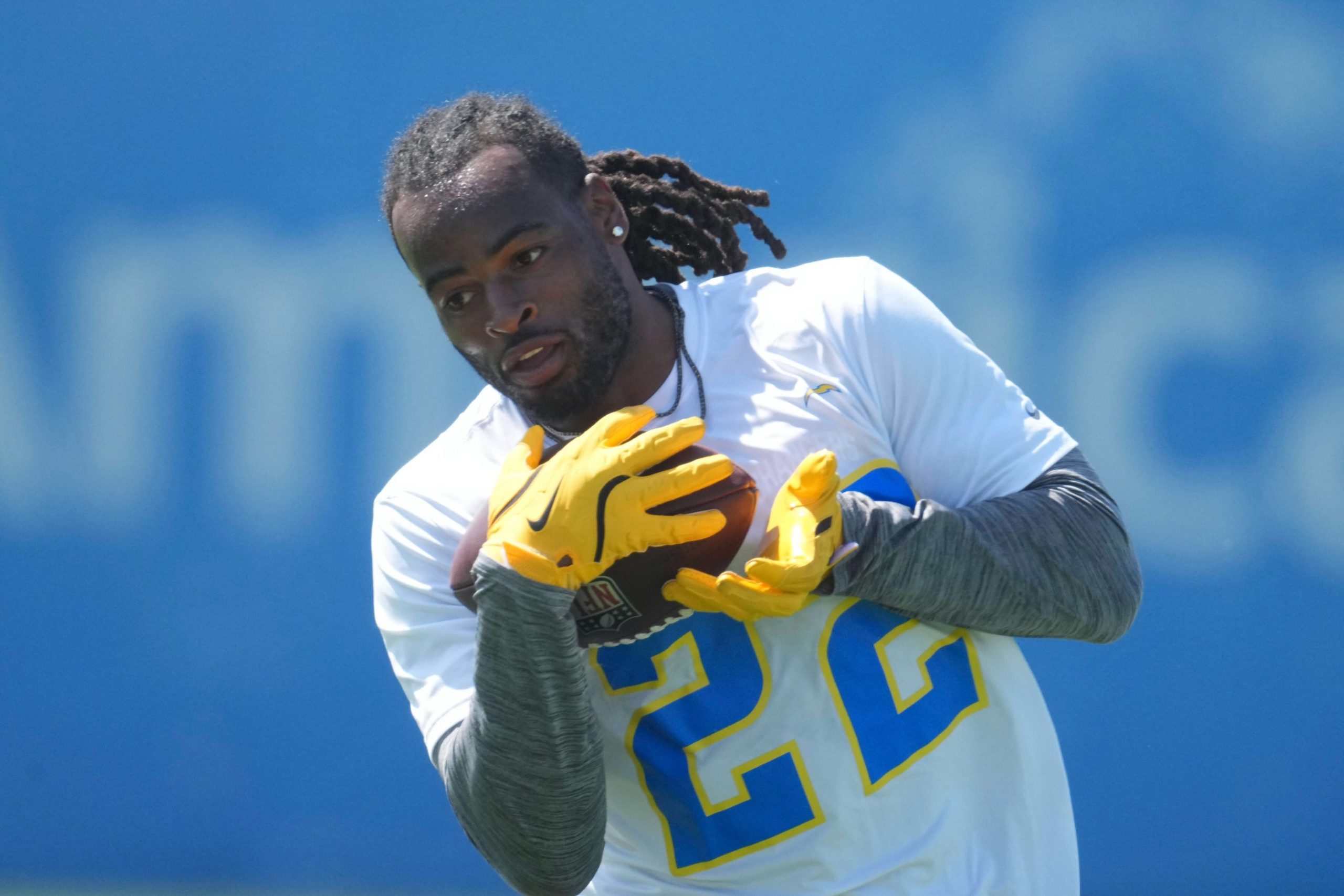 Ein Footballspieler mit Dreadlocks, der Najee Harris ähnelt, trägt ein weißes Trikot mit der Nummer 22 und gelbe Handschuhe und bereitet sich darauf vor, einen Football beim Training zu fangen. Der blaue, leicht verschwommene Hintergrund hebt die Aktion des Sportlers hervor. Diese Beschreibung wurde mit der FootballR KI automatisch generiert.