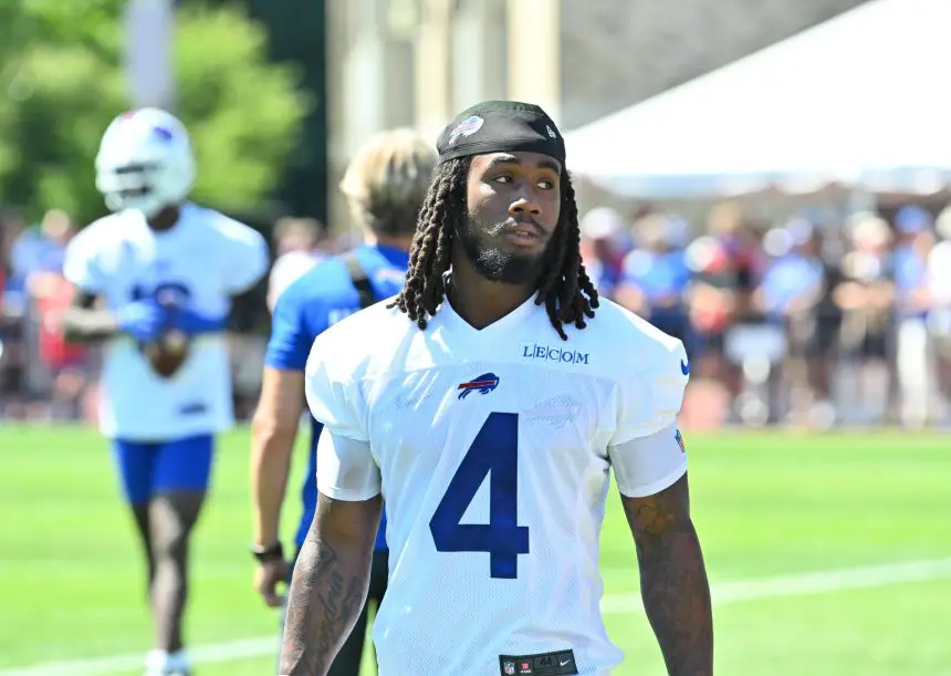 Ein Football-Spieler, der aufgrund seiner weißen Buffalo Bills-Trikotnummer 4 und seiner langen Dreadlocks als James Cook identifiziert werden kann, läuft während des Trainings auf einem grasbewachsenen Feld, während andere Spieler, Mitarbeiter und eine Menschenmenge im Hintergrund verschwimmen. Diese Beschreibung wurde mit der FootballR KI automatisch generiert.