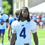 Ein Football-Spieler, der aufgrund seiner weißen Buffalo Bills-Trikotnummer 4 und seiner langen Dreadlocks als James Cook identifiziert werden kann, läuft während des Trainings auf einem grasbewachsenen Feld, während andere Spieler, Mitarbeiter und eine Menschenmenge im Hintergrund verschwimmen. Diese Beschreibung wurde mit der FootballR KI automatisch generiert.