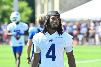 Ein Football-Spieler, der aufgrund seiner weißen Buffalo Bills-Trikotnummer 4 und seiner langen Dreadlocks als James Cook identifiziert werden kann, läuft während des Trainings auf einem grasbewachsenen Feld, während andere Spieler, Mitarbeiter und eine Menschenmenge im Hintergrund verschwimmen. Diese Beschreibung wurde mit der FootballR KI automatisch generiert.