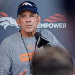 Sean Payton in einem grauen Denver Broncos-Hemd und einer navyfarbenen Broncos-Mütze spricht bei einer Pressekonferenz in ein Mikrofon, während hinter ihm ein Super Bowl-Hintergrund mit dem Broncos-Logo und "EMPOWER" zu sehen ist. Die Szene wirkt offen, mit unscharfen Personen im Vordergrund. Diese Beschreibung wurde mit der FootballR KI automatisch generiert.