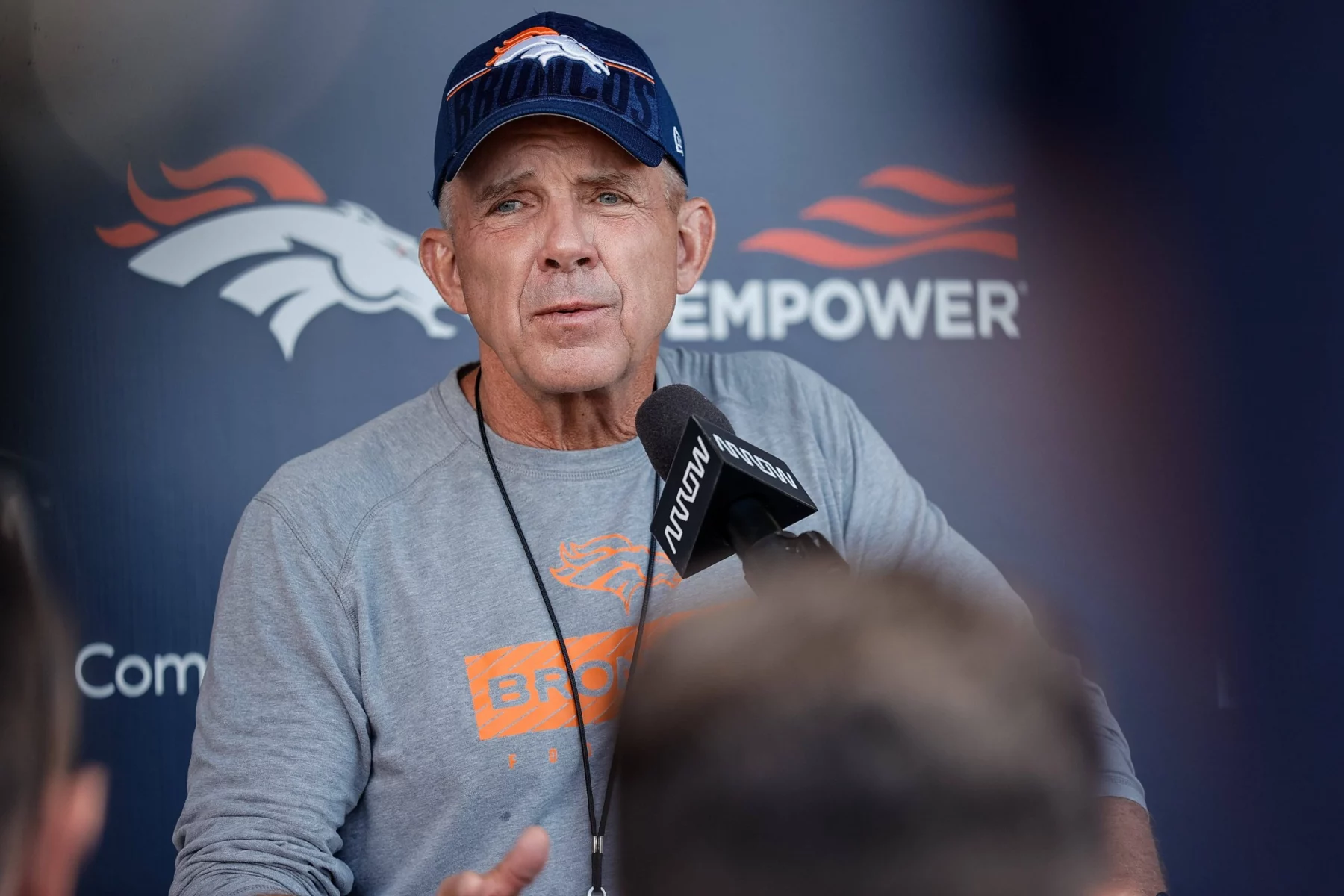 Sean Payton in einem grauen Denver Broncos-Hemd und einer navyfarbenen Broncos-Mütze spricht bei einer Pressekonferenz in ein Mikrofon, während hinter ihm ein Super Bowl-Hintergrund mit dem Broncos-Logo und "EMPOWER" zu sehen ist. Die Szene wirkt offen, mit unscharfen Personen im Vordergrund. Diese Beschreibung wurde mit der FootballR KI automatisch generiert.