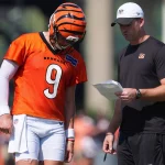 Joe Burrow in einer Bengals-Uniform und einem Helm mit der Nummer 9 steht neben einem Trainer in einem schwarzen Hemd und einer weißen Bengals-Mütze. Der Trainer, möglicherweise Bengals-Streit, hält ein Blatt in der Hand, während sie während des Trainings die Strategie besprechen. Diese Beschreibung wurde mit der FootballR KI automatisch generiert.