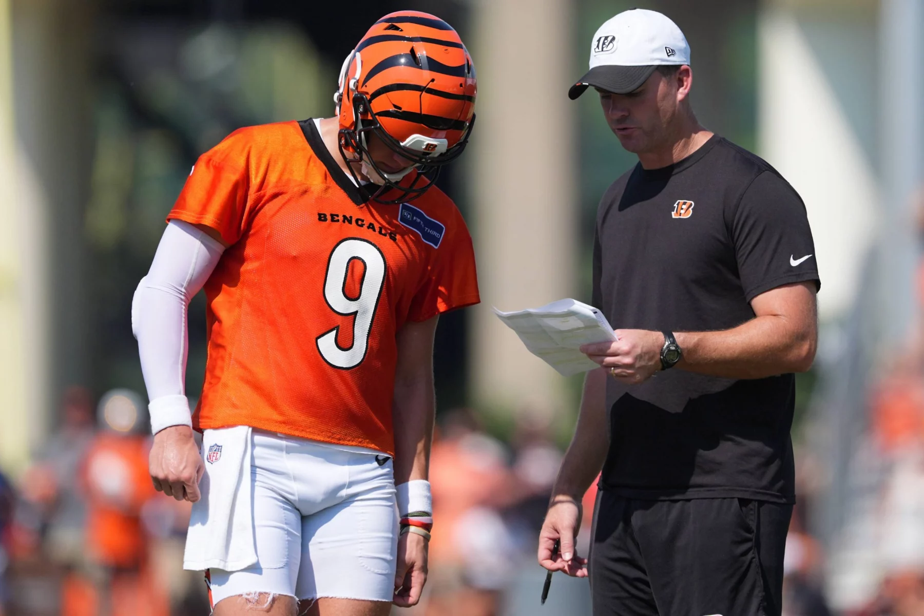 Joe Burrow in einer Bengals-Uniform und einem Helm mit der Nummer 9 steht neben einem Trainer in einem schwarzen Hemd und einer weißen Bengals-Mütze. Der Trainer, möglicherweise Bengals-Streit, hält ein Blatt in der Hand, während sie während des Trainings die Strategie besprechen. Diese Beschreibung wurde mit der FootballR KI automatisch generiert.