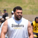 Ein muskulöser Mann mit Bart, der ein ärmelloses weißes "Steelers Football"-Shirt trägt, steht im Freien beim Football-Training - Cameron Heyward und andere in schwarzer und gelber Steelers-Ausrüstung erscheinen in der Nähe eines Grashügels im Hintergrund. Diese Beschreibung wurde mit der FootballR KI automatisch generiert.