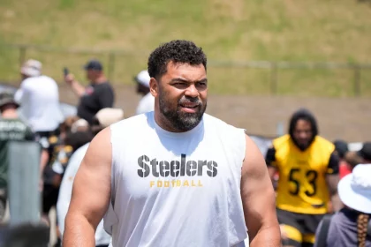 Ein muskulöser Mann mit Bart, der ein ärmelloses weißes "Steelers Football"-Shirt trägt, steht im Freien beim Football-Training - Cameron Heyward und andere in schwarzer und gelber Steelers-Ausrüstung erscheinen in der Nähe eines Grashügels im Hintergrund. Diese Beschreibung wurde mit der FootballR KI automatisch generiert.