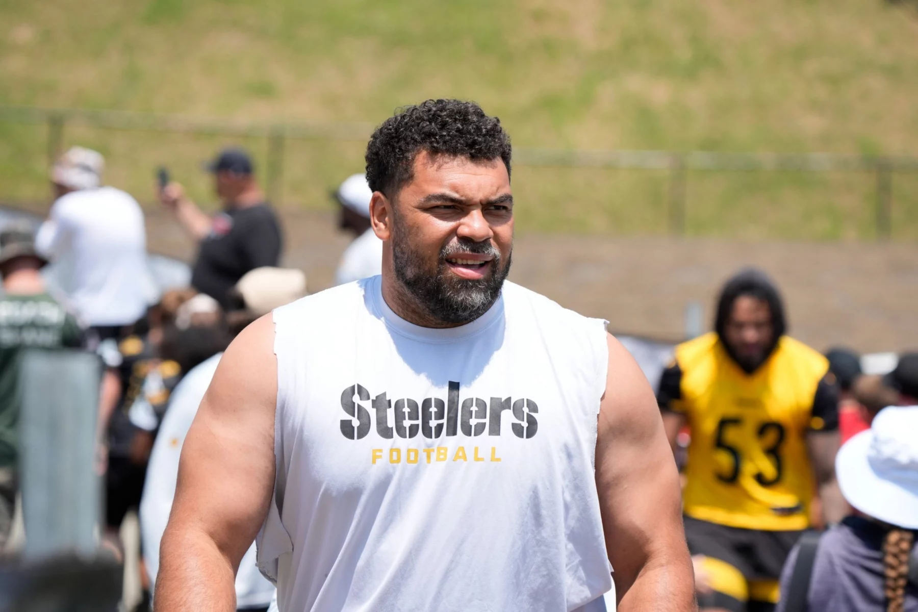 Ein muskulöser Mann mit Bart, der ein ärmelloses weißes "Steelers Football"-Shirt trägt, steht im Freien beim Football-Training - Cameron Heyward und andere in schwarzer und gelber Steelers-Ausrüstung erscheinen in der Nähe eines Grashügels im Hintergrund. Diese Beschreibung wurde mit der FootballR KI automatisch generiert.