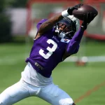 Ein Footballspieler, Jordan Addison, in einem violetten Trikot mit der Nummer 3 streckt beide Arme aus, um einen Football während des Trainings auf einer Wiese zu fangen. Er trägt einen Helm mit Gesichtsschutz und eine weiße Hose. Im Hintergrund sind ein verschwommener Torpfosten und Grünpflanzen zu sehen. Diese Beschreibung wurde mit der FootballR KI automatisch generiert.