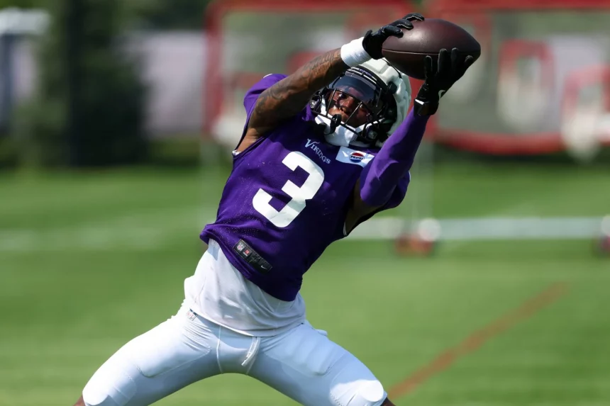 Ein Footballspieler, Jordan Addison, in einem violetten Trikot mit der Nummer 3 streckt beide Arme aus, um einen Football während des Trainings auf einer Wiese zu fangen. Er trägt einen Helm mit Gesichtsschutz und eine weiße Hose. Im Hintergrund sind ein verschwommener Torpfosten und Grünpflanzen zu sehen. Diese Beschreibung wurde mit der FootballR KI automatisch generiert.