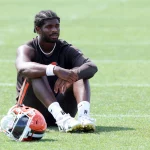 Shedeur Sanders mit geflochtenen Haaren sitzt in Sportkleidung der Cleveland Browns auf dem Rasen, die Arme auf die Knie gestützt. Er trägt ein schwarzes ärmelloses Hemd, schwarze Shorts, weiße Socken und weiße Stollenschuhe. Ein orangefarbener und weißer Helm liegt neben ihm auf dem grünen Feld. Diese Beschreibung wurde mit der FootballR KI automatisch generiert.