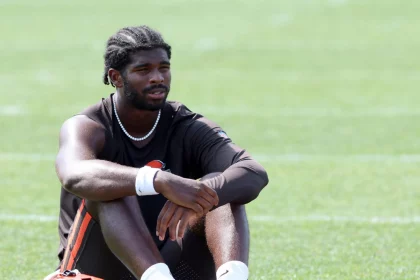 Shedeur Sanders mit geflochtenen Haaren sitzt in Sportkleidung der Cleveland Browns auf dem Rasen, die Arme auf die Knie gestützt. Er trägt ein schwarzes ärmelloses Hemd, schwarze Shorts, weiße Socken und weiße Stollenschuhe. Ein orangefarbener und weißer Helm liegt neben ihm auf dem grünen Feld. Diese Beschreibung wurde mit der FootballR KI automatisch generiert.