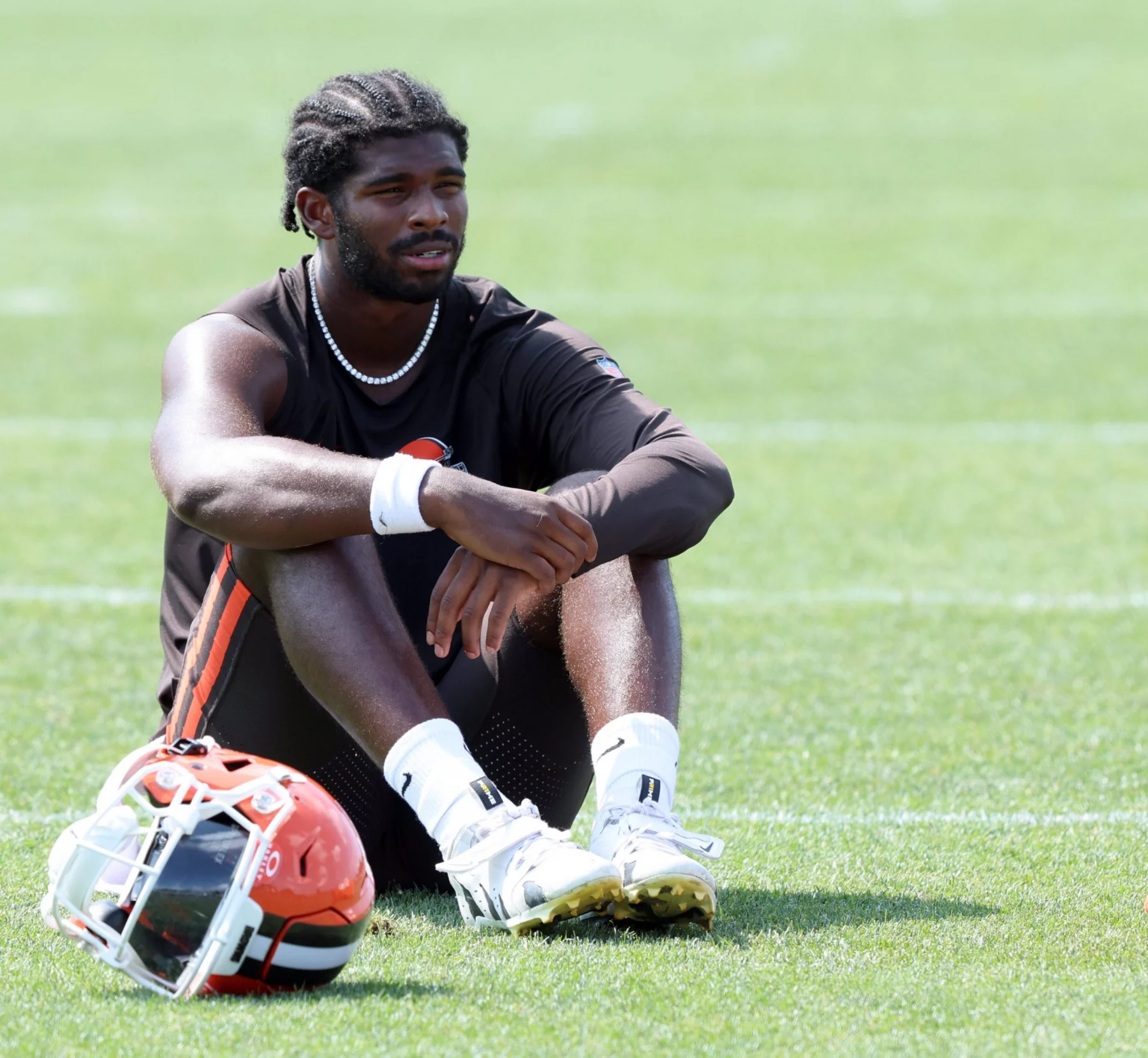 Shedeur Sanders mit geflochtenen Haaren sitzt in Sportkleidung der Cleveland Browns auf dem Rasen, die Arme auf die Knie gestützt. Er trägt ein schwarzes ärmelloses Hemd, schwarze Shorts, weiße Socken und weiße Stollenschuhe. Ein orangefarbener und weißer Helm liegt neben ihm auf dem grünen Feld. Diese Beschreibung wurde mit der FootballR KI automatisch generiert.