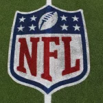 Das NFL-Logo mit dem Football und acht weißen Sternen über dem fetten roten Schriftzug "NFL" ist auf grünem Kunstrasen aufgemalt - ein Symbol, das im Mittelpunkt von Kaderbau und Cap Space Strategien steht, wenn Teams ihren Cap Space für den Erfolg in der Meisterschaft verwalten. Diese Beschreibung wurde mit der FootballR KI automatisch generiert.
