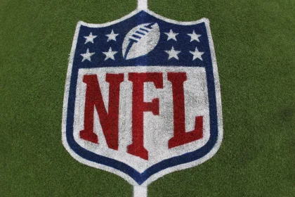 Das NFL-Logo mit dem Football und acht weißen Sternen über dem fetten roten Schriftzug "NFL" ist auf grünem Kunstrasen aufgemalt - ein Symbol, das im Mittelpunkt von Kaderbau und Cap Space Strategien steht, wenn Teams ihren Cap Space für den Erfolg in der Meisterschaft verwalten. Diese Beschreibung wurde mit der FootballR KI automatisch generiert.