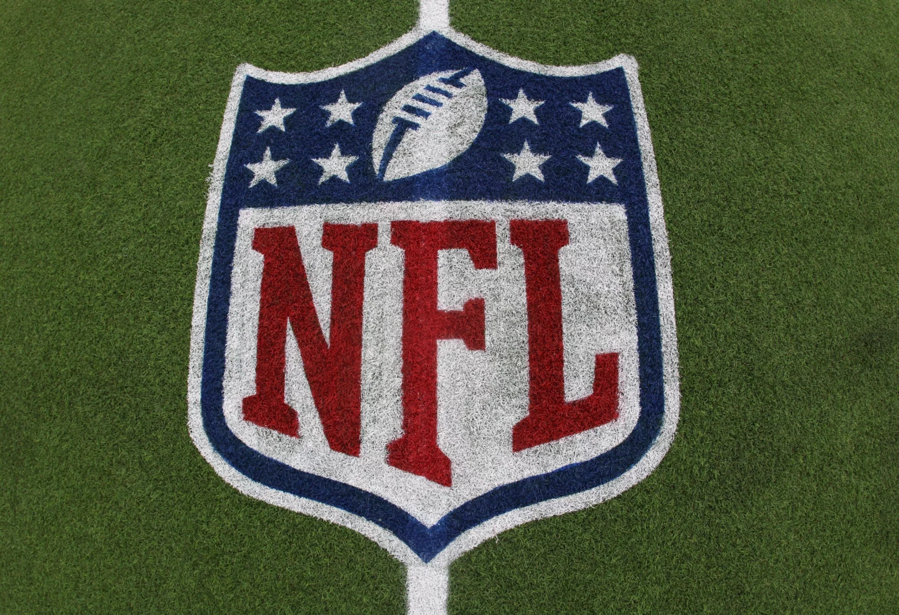 Das NFL-Logo mit dem Football und acht weißen Sternen über dem fetten roten Schriftzug "NFL" ist auf grünem Kunstrasen aufgemalt - ein Symbol, das im Mittelpunkt von Kaderbau und Cap Space Strategien steht, wenn Teams ihren Cap Space für den Erfolg in der Meisterschaft verwalten. Diese Beschreibung wurde mit der FootballR KI automatisch generiert.