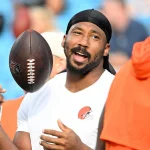 Woche 8 - Ein Mann, der ein weißes Cleveland-Browns-Hemd und eine schwarze Kutte trägt, Myles Garrett, lächelt und spricht, während er einen Football auf seinem Finger dreht. Er hat einen kurzen Bart und Schnurrbart. Andere Personen in orangefarbenen Hemden sind in der Nähe, mit verschwommenen Stadionsitzen hinter ihnen. Diese Beschreibung wurde mit der FootballR KI automatisch generiert.