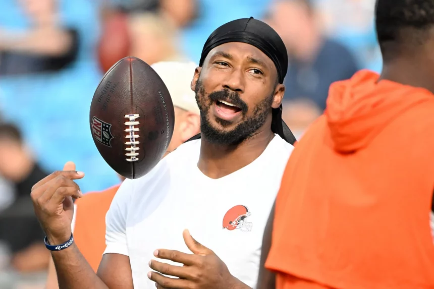 Woche 8 - Ein Mann, der ein weißes Cleveland-Browns-Hemd und eine schwarze Kutte trägt, Myles Garrett, lächelt und spricht, während er einen Football auf seinem Finger dreht. Er hat einen kurzen Bart und Schnurrbart. Andere Personen in orangefarbenen Hemden sind in der Nähe, mit verschwommenen Stadionsitzen hinter ihnen. Diese Beschreibung wurde mit der FootballR KI automatisch generiert.