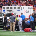 Morice Norris - Eine Gruppe von medizinischem Personal versammelt sich um eine Person, die auf einem Footballfeld vor einem Krankenwagen mit der Aufschrift "Grady" liegt, während im Hintergrund eine Menschenmenge im Stadion zusieht. Notfallausrüstung und Tragen sind auf dem Rasen zu sehen. Diese Beschreibung wurde mit der FootballR KI automatisch generiert.