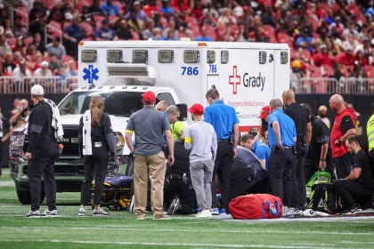 Morice Norris - Eine Gruppe von medizinischem Personal versammelt sich um eine Person, die auf einem Footballfeld vor einem Krankenwagen mit der Aufschrift "Grady" liegt, während im Hintergrund eine Menschenmenge im Stadion zusieht. Notfallausrüstung und Tragen sind auf dem Rasen zu sehen. Diese Beschreibung wurde mit der FootballR KI automatisch generiert.