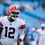 Ein Football-Spieler in einem Trikot der Cleveland Browns, mit der Nummer 12 und einem orangefarbenen Helm, steht auf dem Spielfeld. Er erinnert an Shedeur Sanders, lächelt und blickt nach links, während die Sitze im Stadion hinter ihm weitgehend leer sind. Diese Beschreibung wurde mit der FootballR KI automatisch generiert.