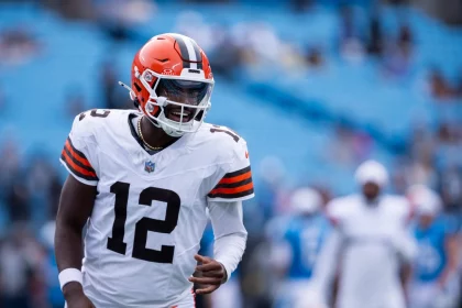 Ein Football-Spieler in einem Trikot der Cleveland Browns, mit der Nummer 12 und einem orangefarbenen Helm, steht auf dem Spielfeld. Er erinnert an Shedeur Sanders, lächelt und blickt nach links, während die Sitze im Stadion hinter ihm weitgehend leer sind. Diese Beschreibung wurde mit der FootballR KI automatisch generiert.