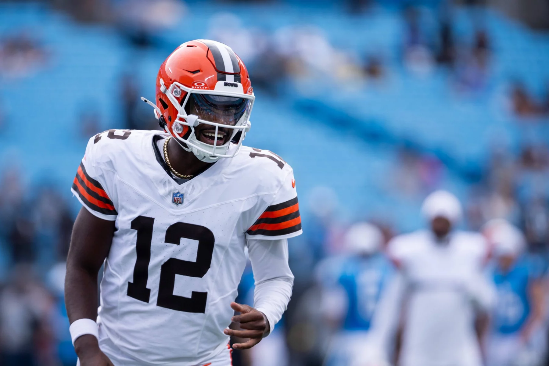 Ein Football-Spieler in einem Trikot der Cleveland Browns, mit der Nummer 12 und einem orangefarbenen Helm, steht auf dem Spielfeld. Er erinnert an Shedeur Sanders, lächelt und blickt nach links, während die Sitze im Stadion hinter ihm weitgehend leer sind. Diese Beschreibung wurde mit der FootballR KI automatisch generiert.