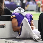 Ein Football-Spieler in einer lila Minnesota Vikings-Uniform liegt auf dem Rücken auf einem Krankenwagen, ein weißes Handtuch bedeckt seinen Kopf. Er scheint verletzt zu sein, ein Arm ruht auf seinem Knie - eine Erinnerung an die Momente, die Rondale Moore erlebt hat. Diese Beschreibung wurde mit der FootballR KI automatisch generiert.