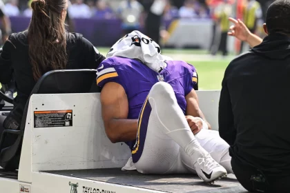 Ein Football-Spieler in einer lila Minnesota Vikings-Uniform liegt auf dem Rücken auf einem Krankenwagen, ein weißes Handtuch bedeckt seinen Kopf. Er scheint verletzt zu sein, ein Arm ruht auf seinem Knie - eine Erinnerung an die Momente, die Rondale Moore erlebt hat. Diese Beschreibung wurde mit der FootballR KI automatisch generiert.