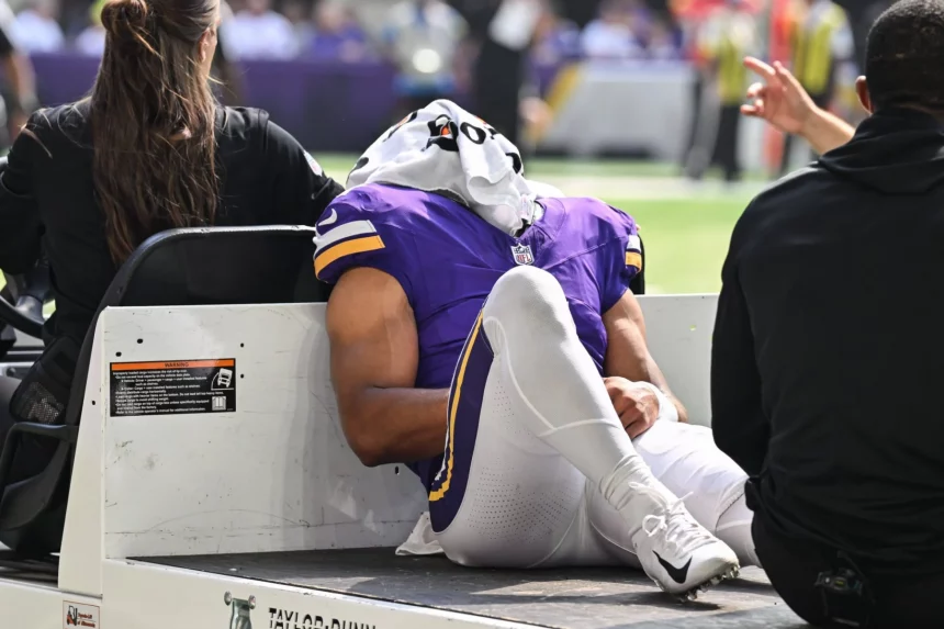 Ein Football-Spieler in einer lila Minnesota Vikings-Uniform liegt auf dem Rücken auf einem Krankenwagen, ein weißes Handtuch bedeckt seinen Kopf. Er scheint verletzt zu sein, ein Arm ruht auf seinem Knie - eine Erinnerung an die Momente, die Rondale Moore erlebt hat. Diese Beschreibung wurde mit der FootballR KI automatisch generiert.